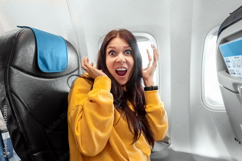 A girl sits in front of an airplane window and is nervous, afraid to fly, the cabin of a passenger airliner. Aerophobia, phobia, international flights, stress, motion sickness. - obrazy, fototapety, plakaty 
