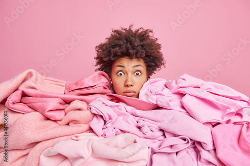 Принти на полотні Shocked young African American woman sticks head through pile of laundry stares stunned at camera busy doing housework collects clothes for washing isolated over pink background