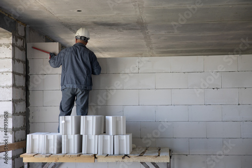 A worker in a hard hat puts a wall of large white bricks. The process of building a house. Construction site.