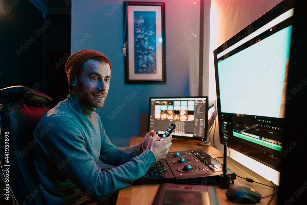 Attractive Male Video Editor Works with Footage or Video on His Personal Computer and having a break communicating on his smartphone. He Works in Creative Office Studio or home. Neon lights