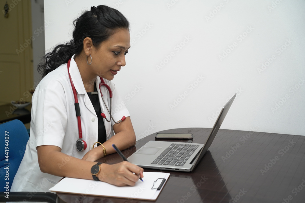 A south Indian female doctor in 30s with laptop and prescription board with white coat and red stethoscope in white background.Online medical consultation A south Indian female doctor in 30s with laptop and prescription board with white coat and red stethoscope in white background.Online medical consultation