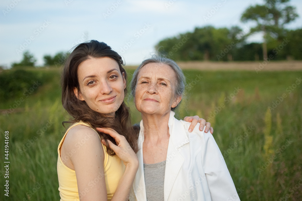 Senior mother with gray hair with her adult daughter looking at the camera in the garden and hugging each other during sunny day outdoors Senior mother with gray hair with her adult daughter looking at the camera in the garden and hugging each other during sunny day outdoors