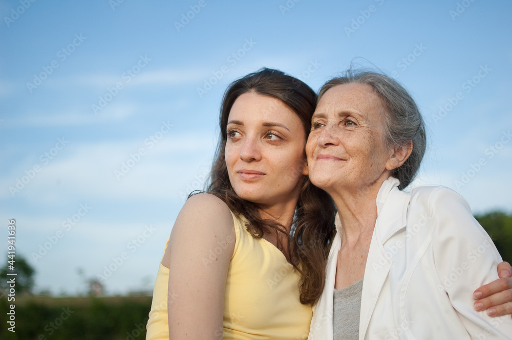 Senior mother with gray hair with her adult daughter looking at the camera in the garden and hugging each other during sunny day outdoors Senior mother with gray hair with her adult daughter looking at the camera in the garden and hugging each other during sunny day outdoors