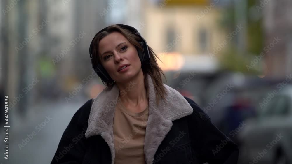 a woman wearing headphones and a jacket with fur trim walks along a city street. blurred background a woman wearing headphones and a jacket with fur trim walks along a city street. blurred background