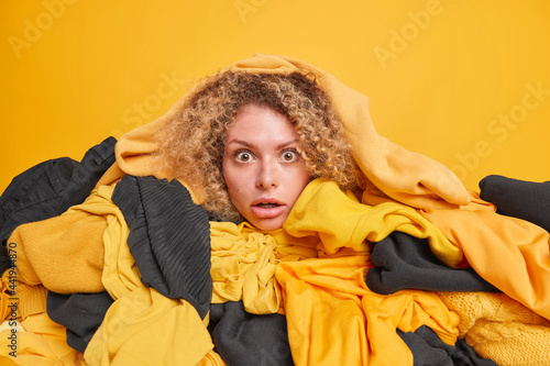 Emotional woman with curly hair chooses what to wear poses among pile of clothes stares impressed at camera isolated over yellow background. Spring cleanig of closet. Sorting and cleaning up. photo