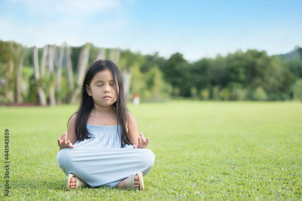 Asian child cute meditate or kid girl close eye mindfulness meditating with breath and sit on lawn or meadow green grass for peace and yoga relax with hand pleat on tree garden at outdoor temple park