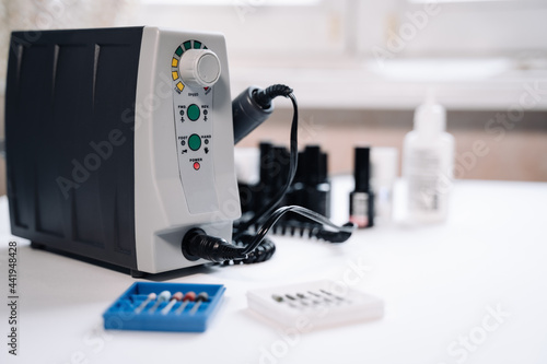 There is a manicure machine on a white table. In the foreground are boxes with attachments (cutters). There are gel polish in the background.