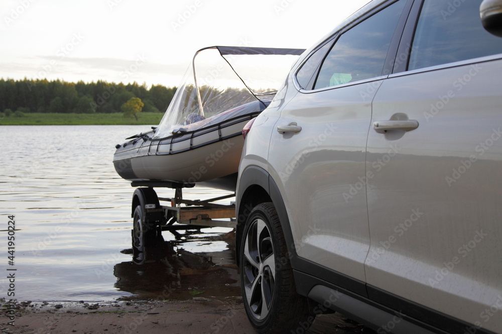A new Korean white SUV crossover car with Russian inflatable boat on trailer in the water on sandy slipway on the river, boat launch before active recreation on the river in Russia, side view closeup