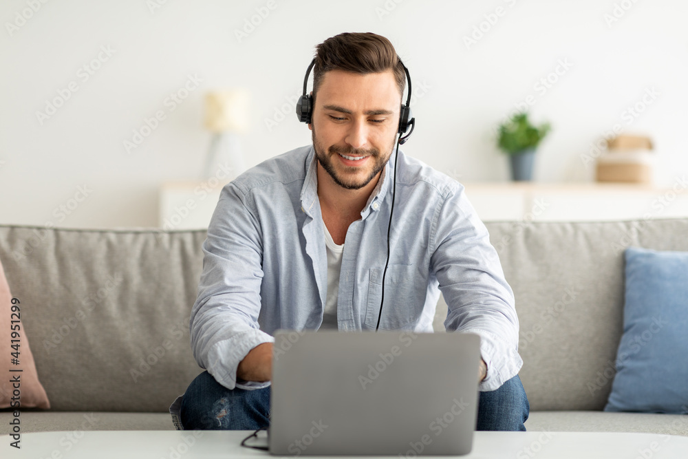 Distance learning concept. Positive man in headset studying online via laptop, sitting on sofa at home, copy space Distance learning concept. Positive man in headset studying online via laptop, sitting on sofa at home, copy space