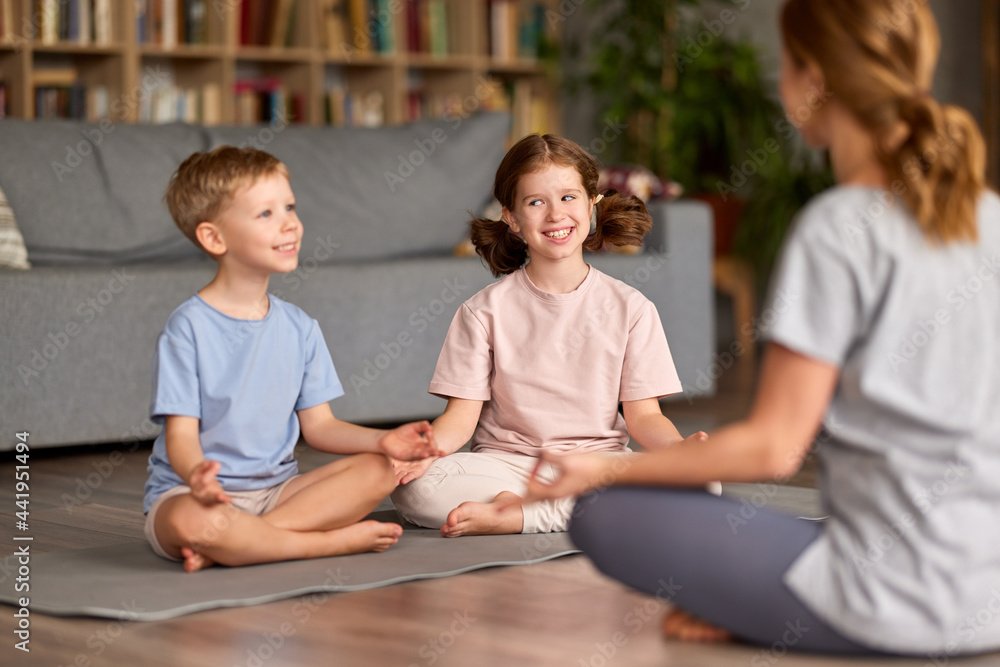 Two happy kids brother and sister in lotus pose keeping smiling and meditating with mom while sitting in living room at home. Mother teaching children son and daughter how to meditate Two happy kids brother and sister in lotus pose keeping smiling and meditating with mom while sitting in living room at home. Mother teaching children son and daughter how to meditate