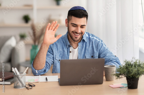 Фотография Smiling israeli man working on laptop waving hand