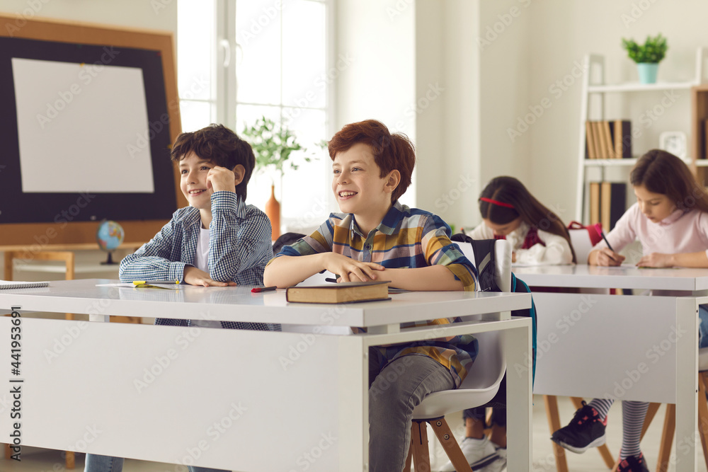 Students in class. Happy children learning new interesting stuff in a lesson. Two happy smiling elementary school boys listening to teacher sitting at desk in modern classroom. Education concept Students in class. Happy children learning new interesting stuff in a lesson. Two happy smiling elementary school boys listening to teacher sitting at desk in modern classroom. Education concept