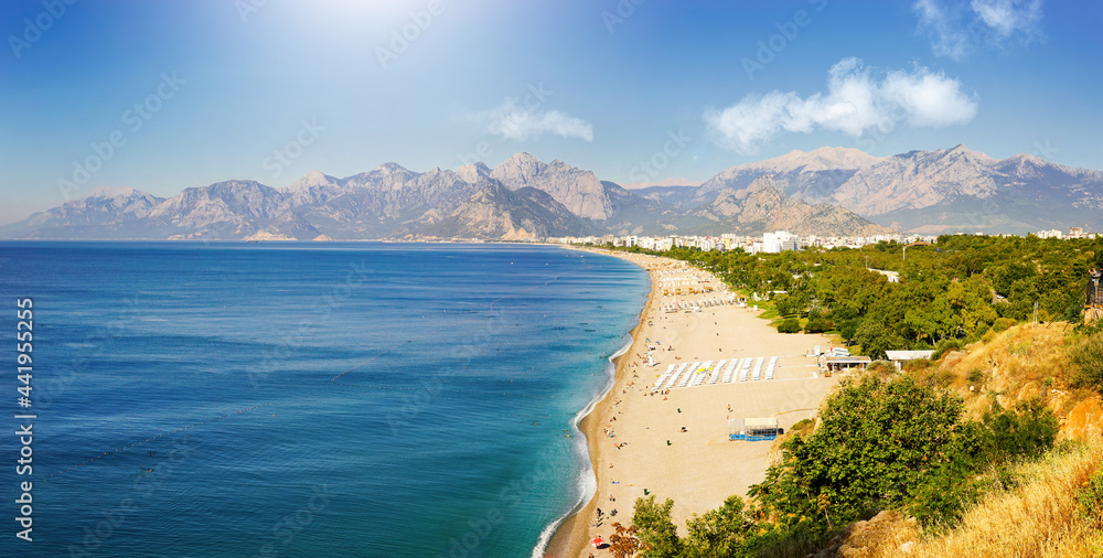 Fototapeta premium Amazingly beautiful panorama of Mediterranean resort Antalya: turquoise sea, white surf line, clean wide beach, greenery of parks, chain of mountains on horizon with clouds, sun and bright blue sky.