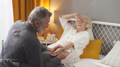 responsive careful senior man check the pulse of sick wife lying on bed at home, help to ill female, touching hand. photo