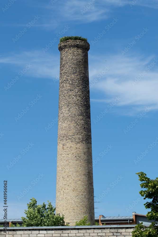 An old vintage brick factory chimney from the industrial revolution days with green grass growing on the top on a cloudy blue summer sky background