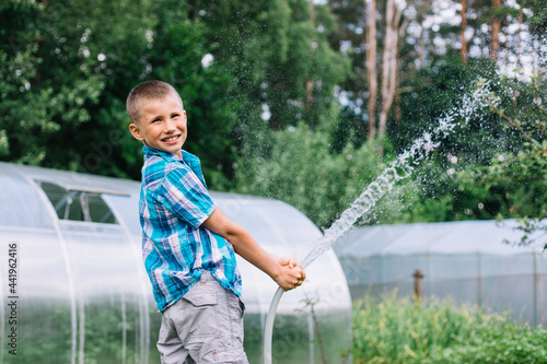 Cute boy in plaid shirt playing with garden sprinkler on summer hot day. Teenager having fun on backyard spraying with a hosepipe. Happy childhood.