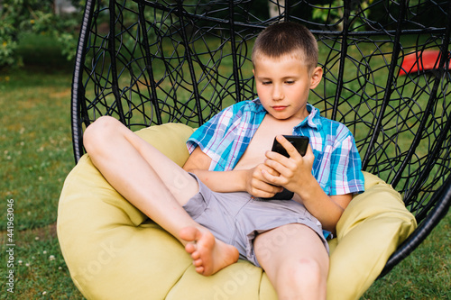 Boy in plaid shirt sitting in cocoon chair and playing with telephone. Smartphone games. Summer time