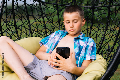 Boy in plaid shirt sitting in cocoon chair and playing with telephone. Smartphone games. Summer time photo