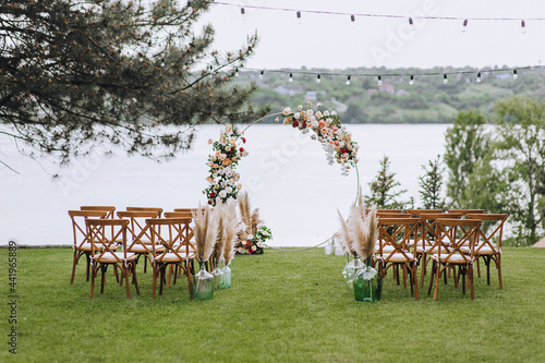 Wedding arch decorated with flowers, wooden chairs stand on green grass against the background of the river on a sunny day. Outdoor ceremony. A place to celebrate. Photography, concept.