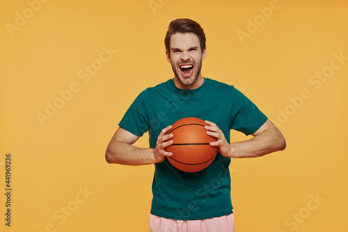 Furious young man holding basketball ball and looking at camera while standing against yellow background
