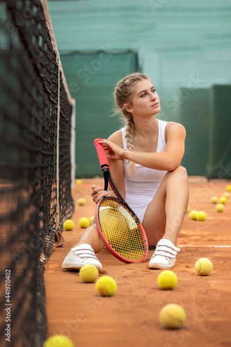 Portrait of beautiful young woman sitting near net in tennis court with ball outdoor. Confident sportswoman resting on tennis court. © volody10