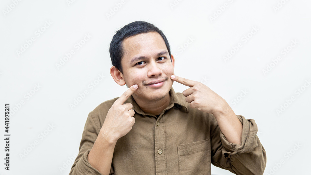 A portrait of young Asian Malay man pointing to his mouth to smile. Keep positive. A portrait of young Asian Malay man pointing to his mouth to smile. Keep positive.