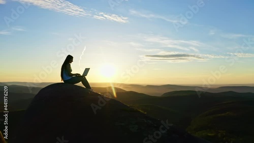 Aerial shots of a silhouette of a young freelance woman working on a laptop sitting on top of a rocky mountain at sunset.