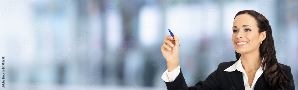Portrait image of happy smiling beautiful businesswoman in black confident suit, writing or drawing something on screen, by marker. Brunette woman, over blurred modern office interior background. Portrait image of happy smiling beautiful businesswoman in black confident suit, writing or drawing something on screen, by marker. Brunette woman, over blurred modern office interior background.