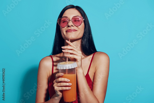 Attractive young woman enjoying cocktail and smiling while standing against blue background