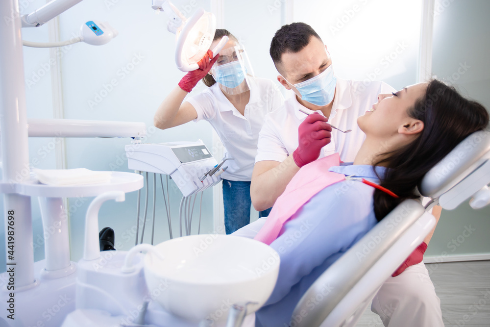 Fototapeta premium Male dentist in protective mask treat the teeth of a young patient. Woman sitting in a dental chair. A young assistant points a lamp at the patient. Medicine during coronavirus quarantine