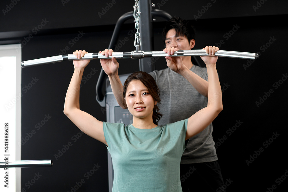 Naklejka premium ラットプルダウンをするアジア人女性と補助をする男性トレーナー