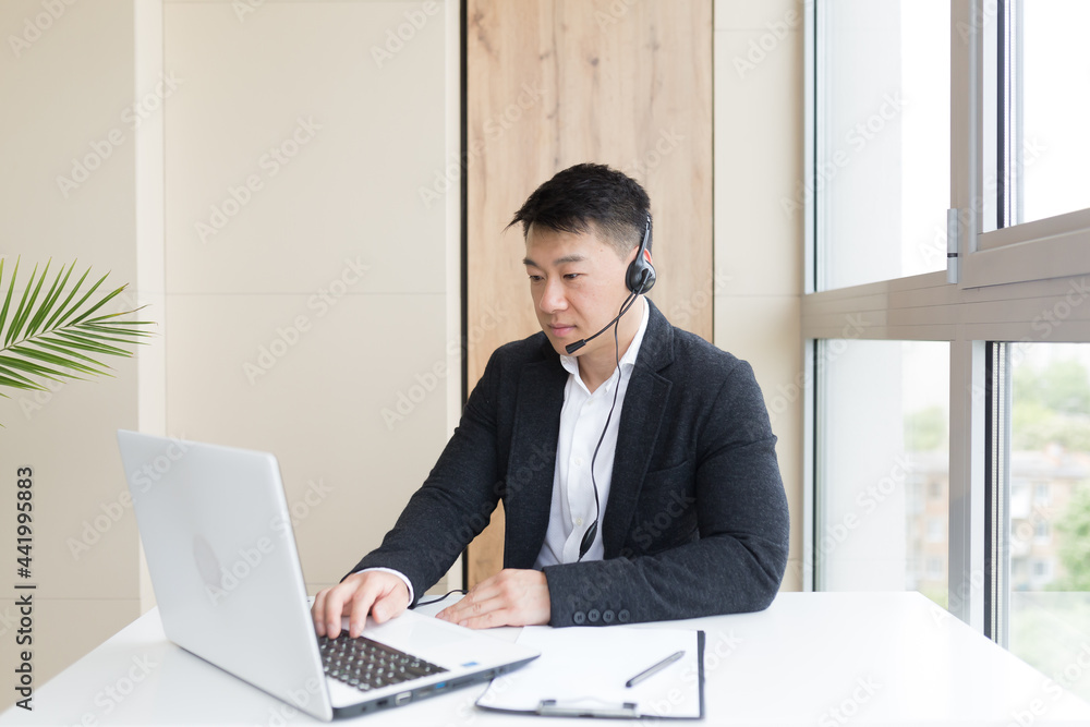 Male businessman conducts online training using a headset and laptop, sitting in a stylish office by the window Male businessman conducts online training using a headset and laptop, sitting in a stylish office by the window