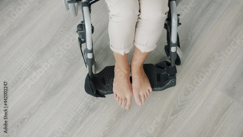 Close-up high angle view of naked female feet in modern wheelchair indoors at home. Disabled paraplegic woman is sitting alone in apartment.