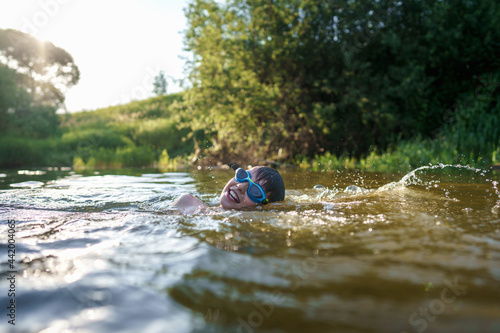Fototapeta Naklejka Na Ścianę i Meble -  Happy boy in swimming glasses swims in the river in the summer at sunset. A child enjoys a summer children's holiday on the shore of the lake. Active holidays. Dynamic Image
