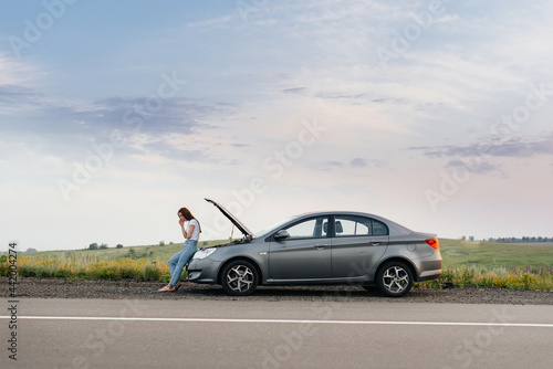 A frustrated young girl stands near a broken-down car in the middle of the highway during sunset. Breakdown and repair of the car. Waiting for help. © Andrii