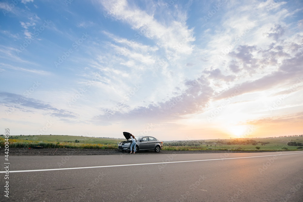 A young girl stands near a broken car in the middle of the highway during sunset and tries to repair it. Breakdown and repair of the car. Troubleshooting the problem. A young girl stands near a broken car in the middle of the highway during sunset and tries to repair it. Breakdown and repair of the car. Troubleshooting the problem.
