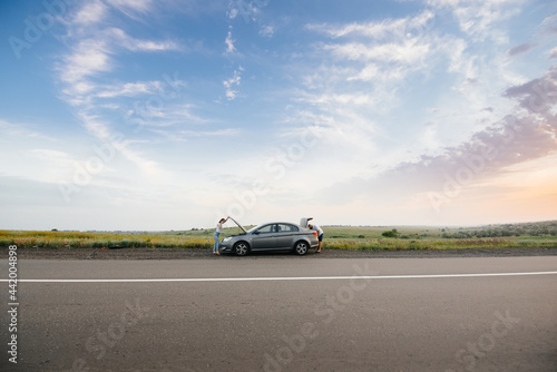 A young girl stands near a broken car in the middle of the highway during sunset and tries to repair it. Breakdown and repair of the car. Troubleshooting the problem. © Andrii