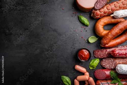 Set of different types of sausages, salami and smoked meat with basil and spices on a black stone background. Top view.