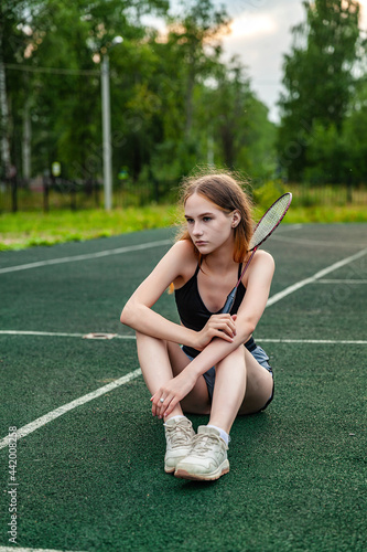 Young beautiful girl in sports top and shorts sitting on the sports ground. Daytime, fitness, open air, badminton, racket