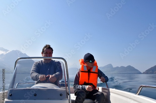 Father and son making a trip on motorboat on lake Lucerne in Switzerland. Boy is wearing orange life jacket and sun glasses. Man is at steering wheel. On background there are snow-capped Swiss Alps. 