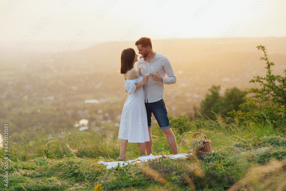 Young elegant couple is having on the hill on a sunny summer day. They are hugging and clinking glasses.
