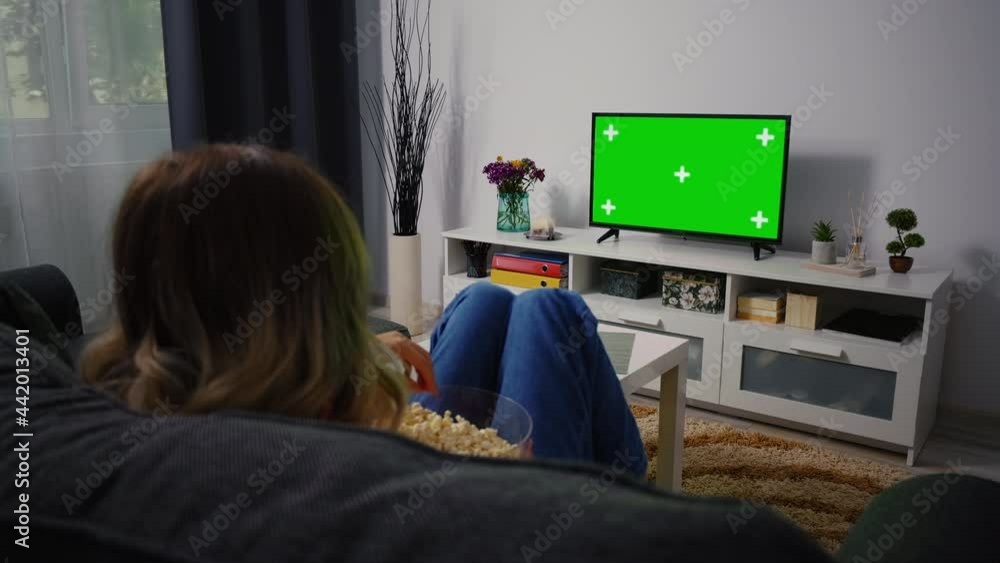 Woman Sitting on a Couch Home Watching Green Chroma Key Screen, Relaxing. Girl in a Cozy Room Watching Sports Match, News, Sitcom TV Show or a Movie on Green Screen eating popcorn. Woman Sitting on a Couch Home Watching Green Chroma Key Screen, Relaxing. Girl in a Cozy Room Watching Sports Match, News, Sitcom TV Show or a Movie on Green Screen eating popcorn.