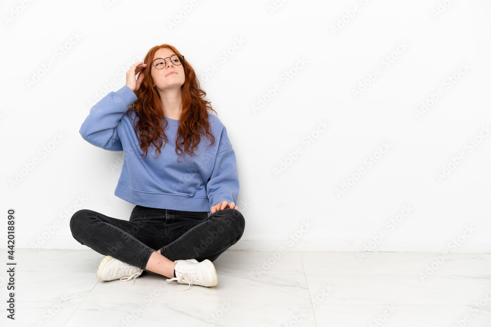 Teenager redhead girl sitting on the floor isolated on white background having doubts and with confuse face expression Teenager redhead girl sitting on the floor isolated on white background having doubts and with confuse face expression