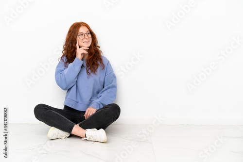 Teenager redhead girl sitting on the floor isolated on white background thinking an idea while looking up
