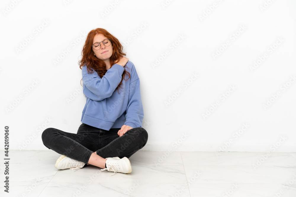 Teenager redhead girl sitting on the floor isolated on white background suffering from pain in shoulder for having made an effort