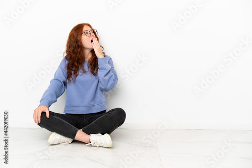 Teenager redhead girl sitting on the floor isolated on white background yawning and covering wide open mouth with hand