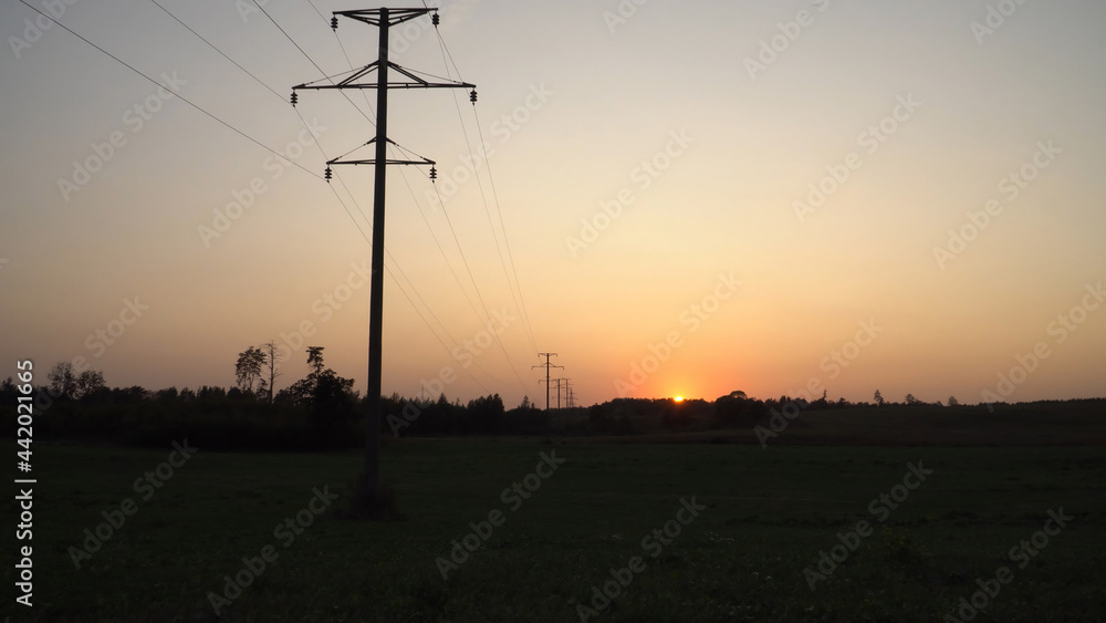 Obraz premium The high voltage power line on sunset time. Landscape in the evening overlooking silhouettes of the electric supply poles in a plain with orange to the golden hue sky background. A view in perspective