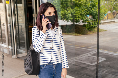 Horizontal portrait of a successful young businesswoman wearing a COVID or coronavirus mask talking about business on her cell phone while walking. Telecommunications concept.