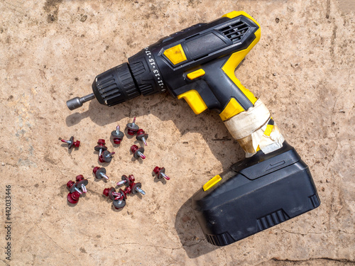 фотографія Black and yellow cordless screwdriver lies on the gray concrete floor next to the crimson short roofing screws