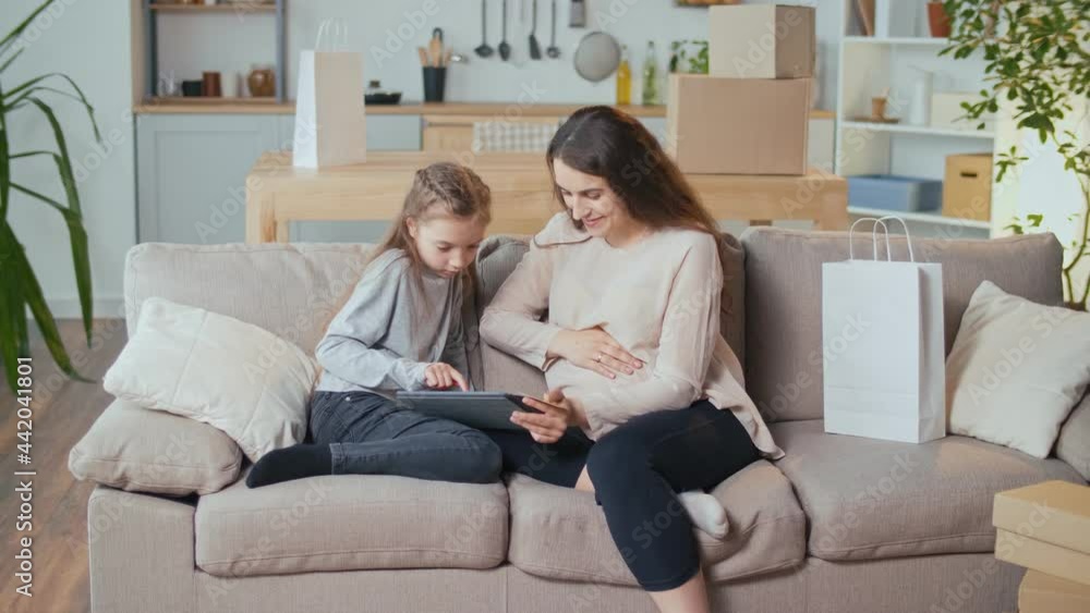 A Pregnant Woman with a Child Makes Online Purchases Using a Computer in a Living Room. Online Delivery Ordering Concept. A Pregnant Woman with a Child Makes Online Purchases Using a Computer in a Living Room. Online Delivery Ordering Concept.
